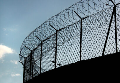 Siluetti vankilan verkkoaidasta, jossa päällä piikkilankaa. Taustalla sininen taivas.