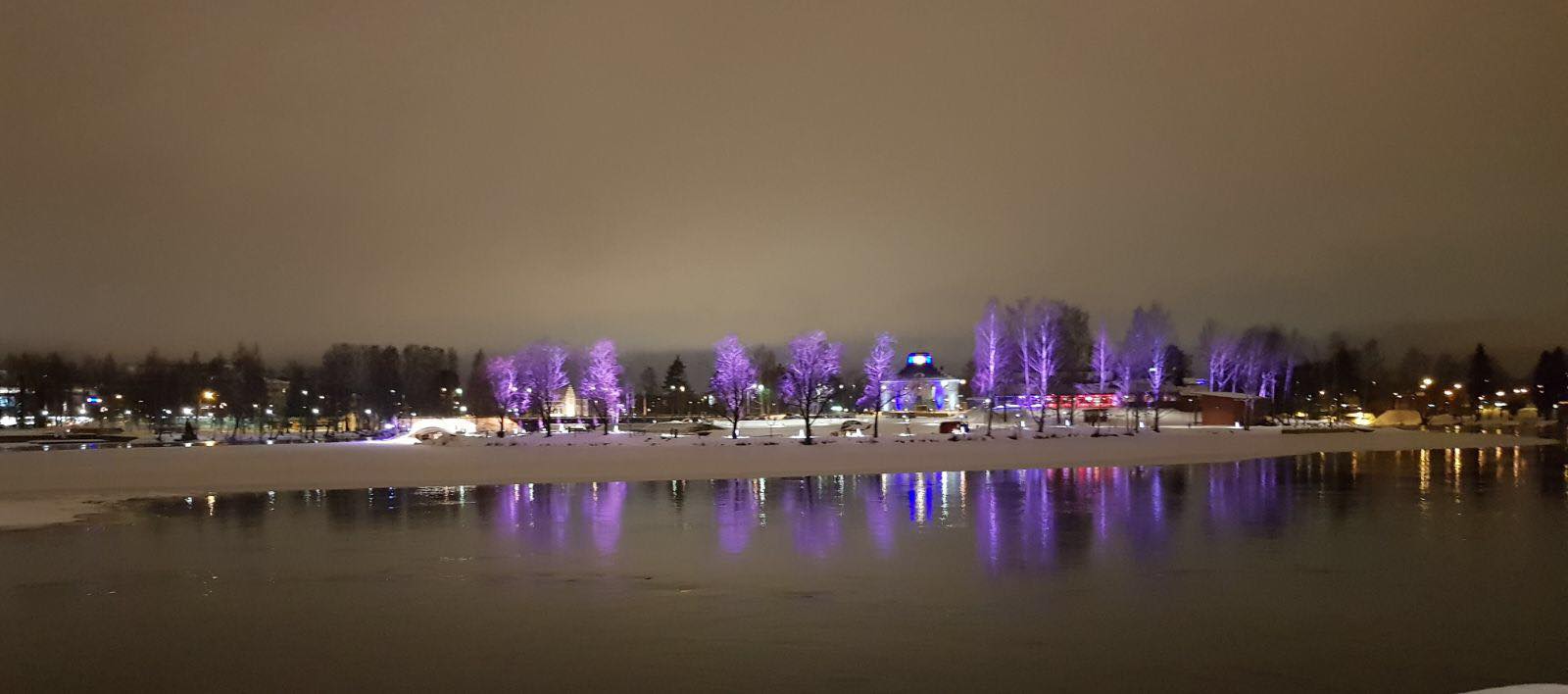 Joen ranta talvella. Vastarannalla lumisia puita ja katuvaloja.