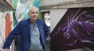 Vaaleahiuksinen mies sinisessä puvussa seisoo graffitiseinän edessä.