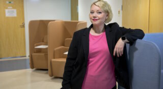 Vaaleahiuksinen nainen seisoo ja nojaa sohvaan