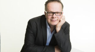 Silmälasipäinen mies puvun takki päällä nojaa pöytään