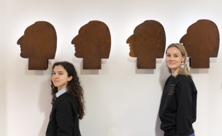 Kaksi naista seinän edessä missä pään muotoisia tauluja