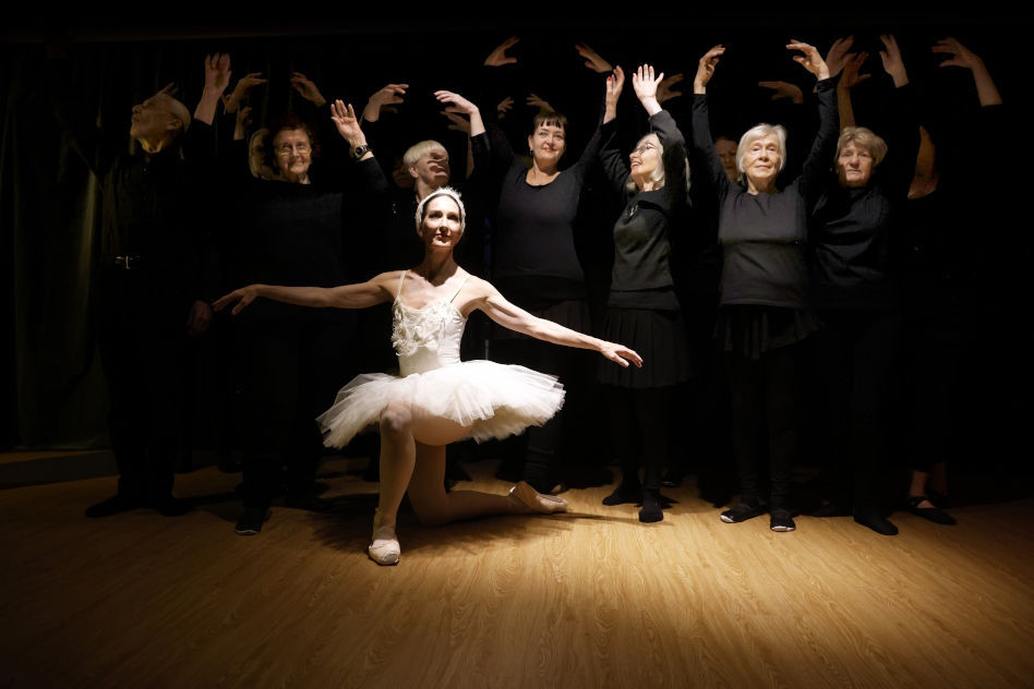 Joukko vanhempia ihmisiä tanssii balettia mustissa vaatteissa, etualalla ballerina vaaleanpunaisessa asussa.