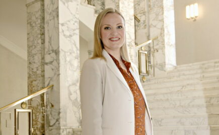 Vaaleahiuksinen nainen valkoisessa jakkupuvussa seisoo eduskuntatalon sisällä, portaiden edessä