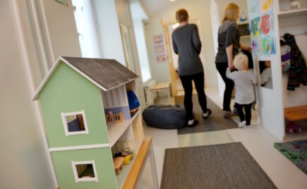 Kaksi aikuista ja lapsi päiväkodissa nukkekodin edessä