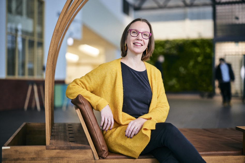 Silmälasipäinen nainen istuu, päällä keltainen neuleliivi