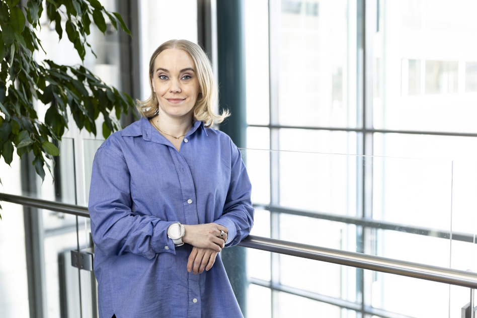 Vaaleahiuksinen nainen seisoo lasiseinän edessä sinisessä puserossa.