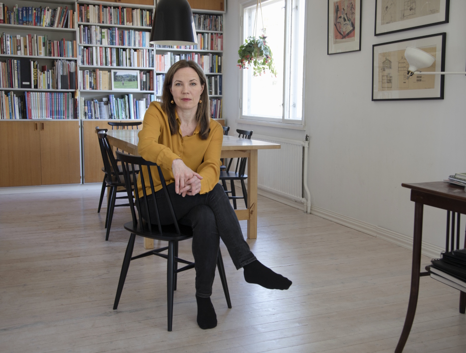 Pitkähiuksinen nainen istuu tuolilla kirjahyllyn edessä