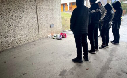 Kolme mustiin pukeutunutta huppupäistä ihmistä seisoo kukkaseinän edessä.