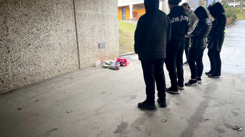 Kolme mustiin pukeutunutta huppupäistä ihmistä seisoo kukkaseinän edessä.