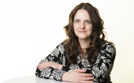 Pitkähiuksinen nainen nojaa pöytään kädet ristissä