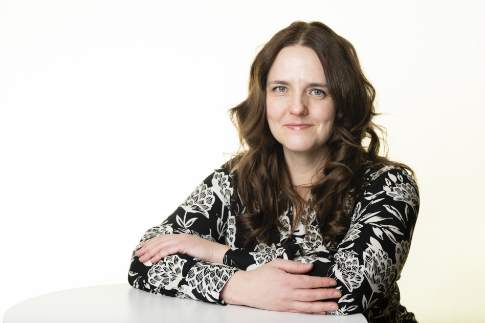 Pitkähiuksinen nainen nojaa pöytään kädet ristissä