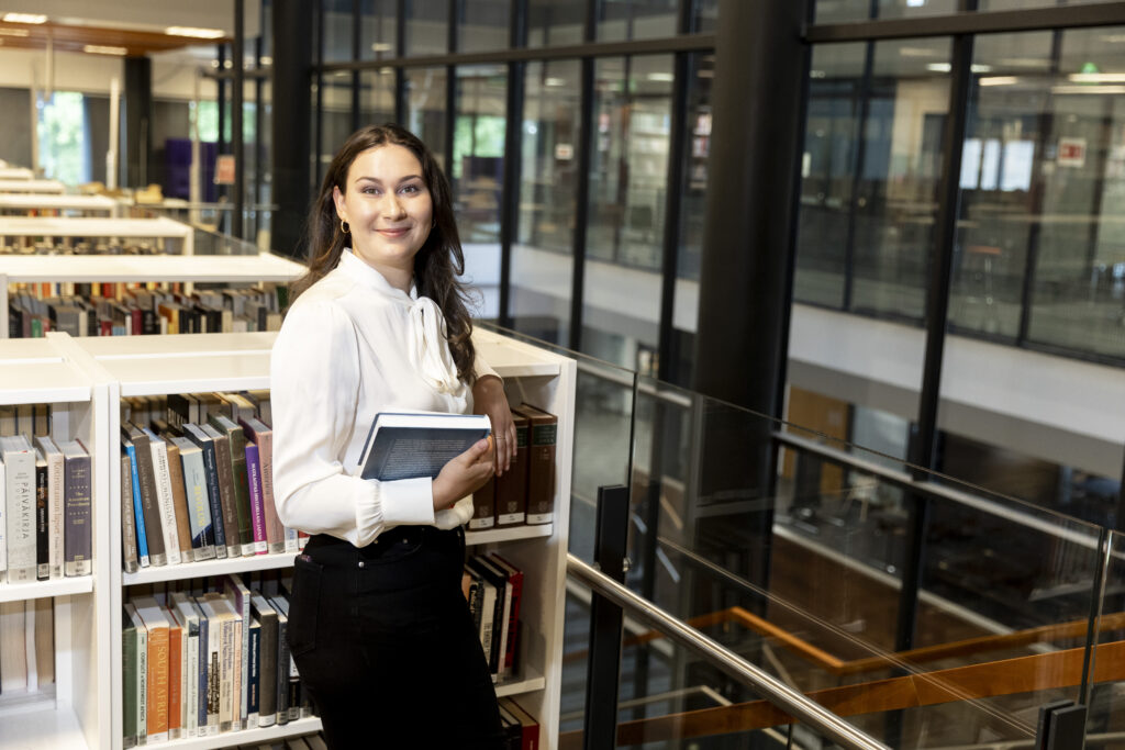 Pitkähiuksinen nainee seisoo kirjastossa kirja kädessään