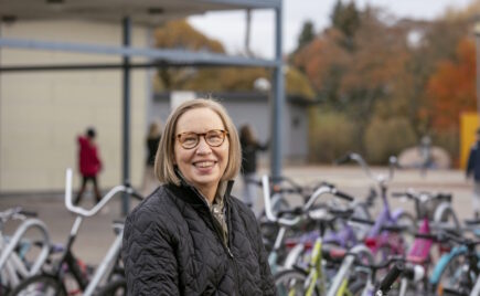 Silmälasipäinen vaaleahiuksinen nainen koulun pihalla pyörätelineen edessä