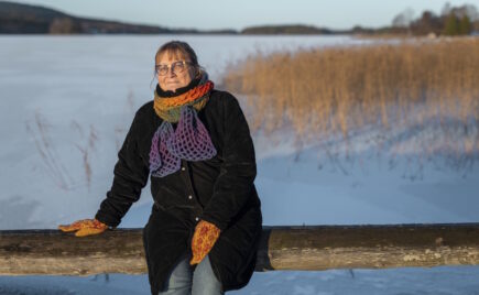 Nainen istuu puunrungon päällä järven rannassa talvimaisemassa.