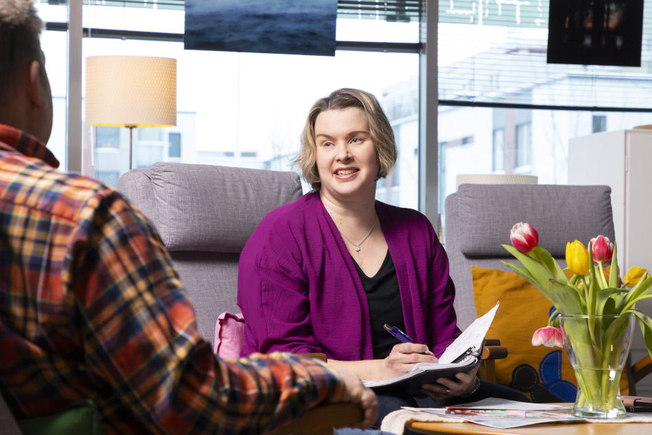 Nainen istuu sohvalla vihko kädessä vastapäätä toista henkilöä.