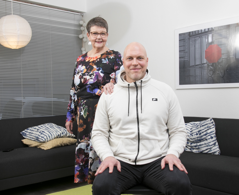 Kalju mies vaaleassa hupparissa istuu etualalla, vanhempi nainen seisoo takana.