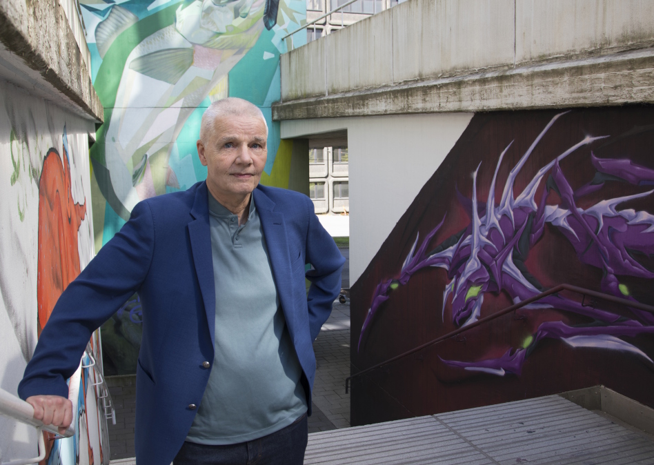 Vaaleahiuksinen mies sinisessä puvussa seisoo graffitiseinän edessä.