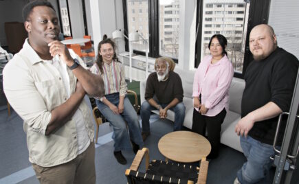 Kolme miestä ja kaksi naista istuu ja seisoo lasiseinäisessä toimistossa.