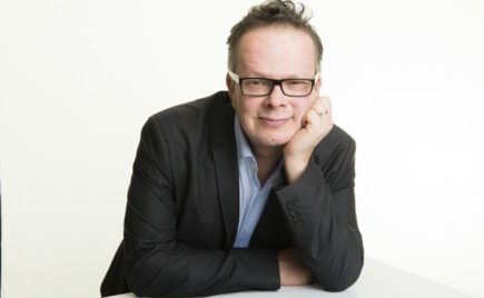 Silmälasipäinen mies puvun takki päällä nojaa pöytään