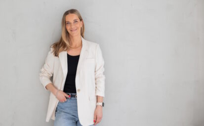 Pitkäniuksinen nuori nainen valkoisessa takissa nojaa seinään. Pauliina Taipelus