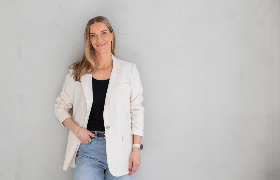 Pitkäniuksinen nuori nainen valkoisessa takissa nojaa seinään. Pauliina Taipelus