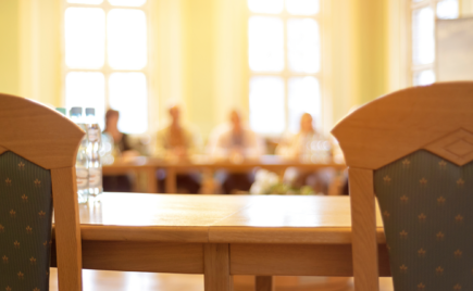 Kokouspöytä, jonka edessä kaksi tyhjää tuolia. Taustalla 4 henkilöä sumennettuina.