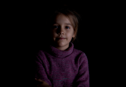 mustalla taustalla ruskeahiuksinen lapsi katsoo kameraan hämärässä valossa