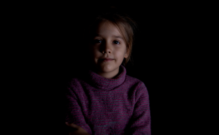 mustalla taustalla ruskeahiuksinen lapsi katsoo kameraan hämärässä valossa