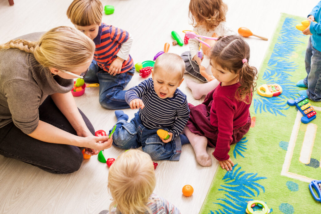 Kuvassa päiväkodin aikuinen ja viisi lasta, jotka leikkivät lattialla. Yhtä lapsista harmittaa ja hänellä on surullinen ilme.