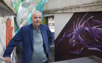 Vaaleahiuksinen mies sinisessä puvussa seisoo graffitiseinän edessä.