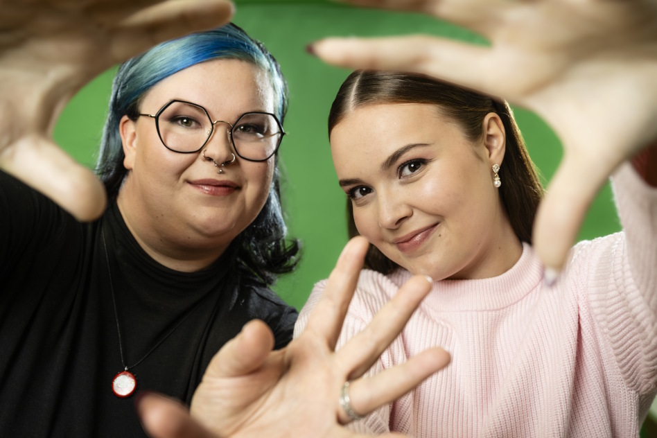 Kaksi nuorta naista vierekkäin, jotka tekevät sormilla sydämen